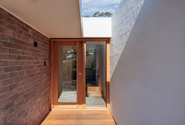 A narrow, sunlit architectural passage leading to a timber-framed glazed door. Exposed red brick forms the left wall, while a tall textured white rendered wall encloses the right side. Timber decking continues through the entry, and an overhead opening brings in daylight and a glimpse of sky and planting. The composition has a clean contemporary character with strong material contrast between brick, warm timber, glass, and white render.