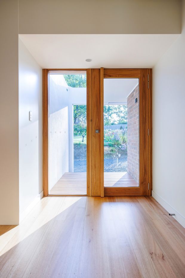 Bright minimalist entryway with pale timber flooring, white walls and ceiling, and full-height natural timber-framed glazed doors opening to a small outdoor courtyard or deck. The simple recessed ceiling light and clean-lined detailing create a calm, contemporary residential interior.