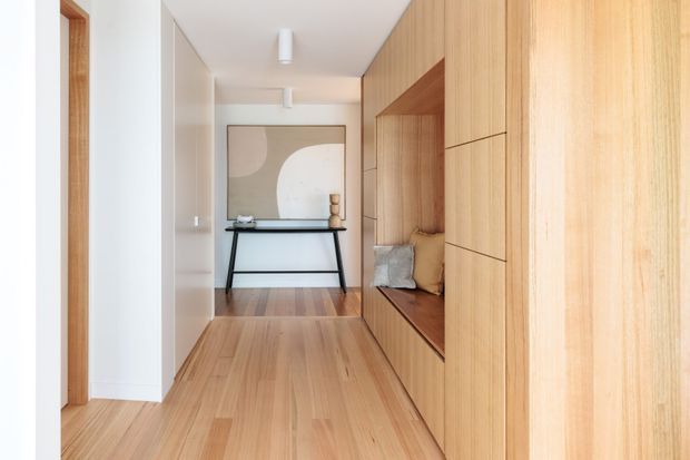 A bright, narrow residential hallway with continuous light timber flooring and extensive built-in timber cabinetry along the right wall. The joinery incorporates an open recessed bench seat with cushions, while a slim black console table sits against the far wall beneath a large neutral abstract artwork. White walls and ceiling, timber-framed door openings, and simple linear ceiling lights create a clean, warm, contemporary interior.