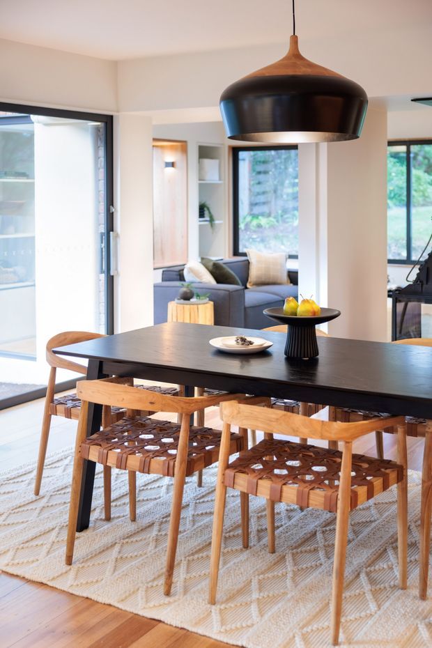 Bright, contemporary open-plan dining area with a long dark dining table, sculptural black pendant light, and timber dining chairs with woven leather-style seats. The room connects visually to a relaxed living area and garden through large windows and glazed doors. White walls, timber flooring, and a pale patterned rug create a warm, airy interior.