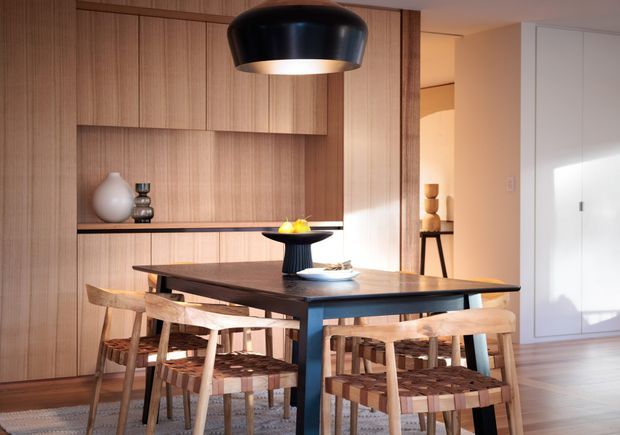 Warm contemporary dining area with a substantial dark dining table surrounded by woven-seat timber chairs. A full-height light timber wall incorporates recessed open shelving and concealed cabinetry, while a large black pendant light hangs above the table. Minimal ceramic and sculptural decor, pale flooring, and a bright adjoining passage create a refined, inviting atmosphere.