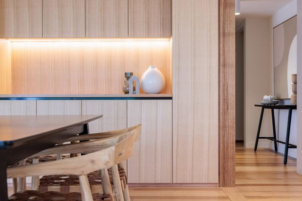 Warm, refined dining area with extensive light timber wall panelling and integrated cabinetry, illuminated by a concealed linear light beneath the upper cupboards. A dark stone or dark-toned benchtop-like ledge runs along the joinery, with a timber dining table and sculptural timber chairs in the foreground. A timber-framed opening leads to a softly styled hallway or adjacent living area with timber flooring and a narrow console table.