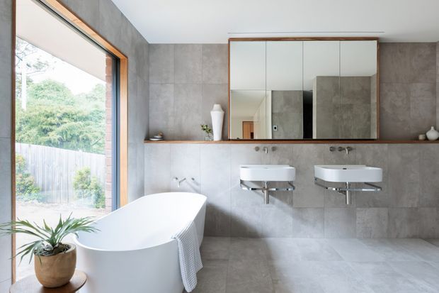 A bright contemporary bathroom with a freestanding white bathtub beside a large timber-framed window, two wall-mounted white basins, and a broad mirrored cabinet above a slim timber shelf. Large grey concrete-look tiles cover the walls and floor, while warm timber framing and sparse ceramic accessories add contrast. Garden greenery is visible through the window.