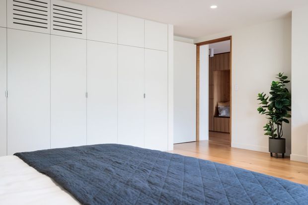 Bright, minimalist bedroom with a low-profile bed in the foreground, a large wall of white built-in wardrobes with ventilation grilles, pale timber flooring, and a timber-framed doorway leading to a darker wood-lined adjoining space. A potted plant adds greenery, while recessed ceiling lighting keeps the room evenly illuminated.