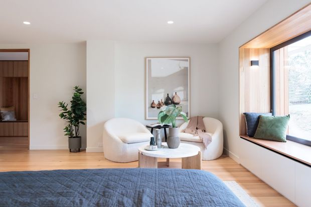 A bright, calm bedroom with a low-profile upholstered bed in the foreground, a small seating area with rounded cream lounge chairs, a round coffee table, indoor plants, framed artwork, and a large timber-framed window with a built-in window seat. The room has pale walls, light timber flooring, recessed ceiling lights, and a warm contemporary fit-out.