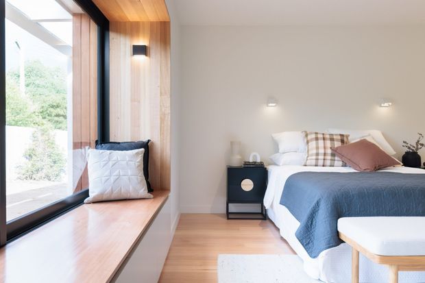 Bright, contemporary bedroom with a large floor-to-ceiling window and integrated timber window seat, pale timber flooring, white walls, and a neatly styled bed with blue bedding and layered cushions. Built-in timber wall and ceiling detailing around the window adds warmth, while compact black bedside furniture and simple wall lights provide contrast.