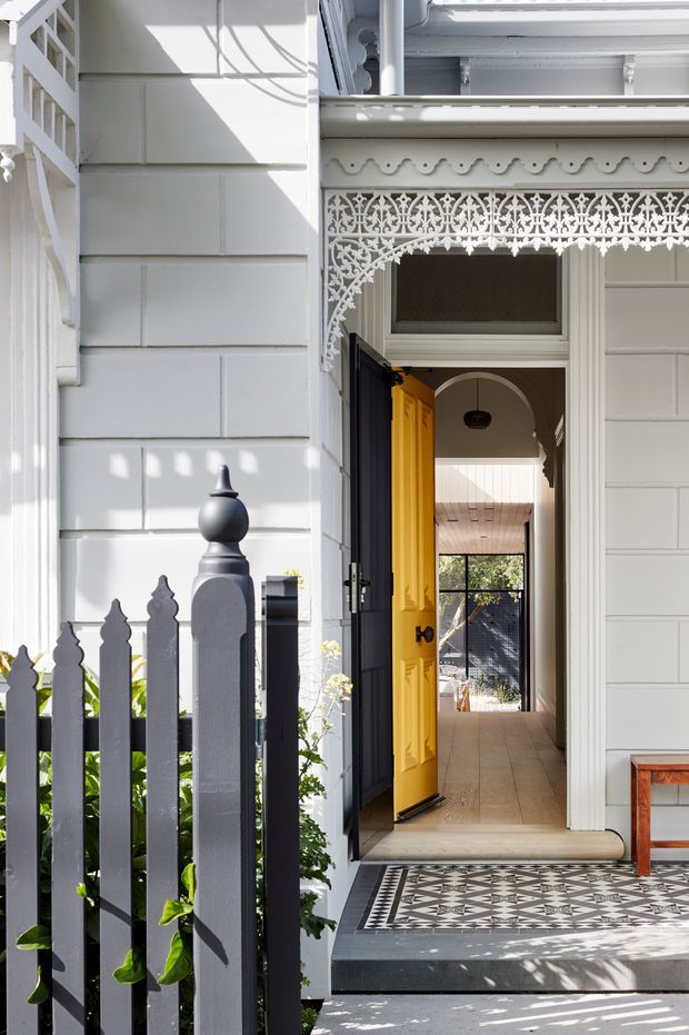 A bright heritage-style residential entrance viewed from outside. The white weatherboard facade features ornate lacework, decorative trim, fluted columns, and a traditional picket fence. A vivid yellow front door opens into a long, tiled hallway, with patterned black-and-white entry tiles and a small timber bench visible at the side.