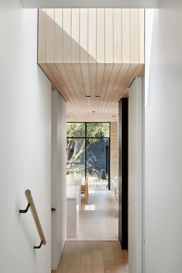 A minimalist interior hallway leading toward a bright living area and glazed courtyard or outdoor space. White walls and built-in doors frame a light timber floor, while the ceiling transitions into a striking continuous pale timber-clad soffit with linear recessed lights. A simple timber handrail is mounted to the left wall, and glimpses of dining and lounge furniture are visible beyond.