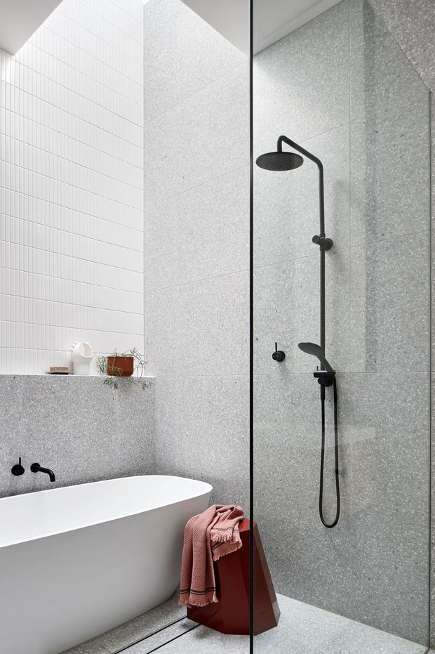 A contemporary bathroom featuring a freestanding white bathtub beside a frameless glass shower enclosure. The walls and floor are clad in light grey speckled stone-look slabs, with a white vertically ribbed tile wall, matte black shower and tapware, and a recessed shelf with small decorative items. A muted terracotta-pink towel adds a soft accent.