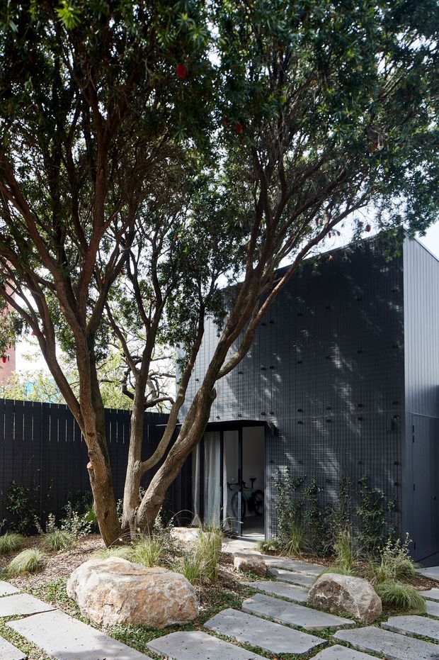 Exterior view of a contemporary dark-clad building partly screened by mature trees. A landscaped entry garden features large natural boulders, ornamental grasses, low planting, and irregular concrete stepping stones leading to a glazed doorway. The building has a tall, simple pitched roof form with vertically ribbed dark cladding and a contrasting lighter side wall.