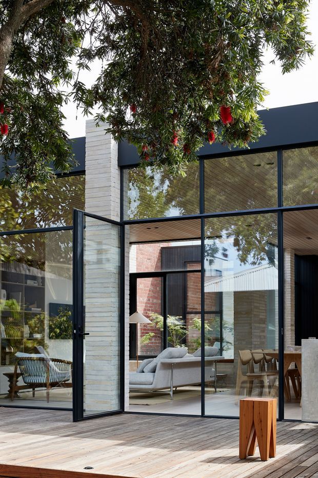 A contemporary glass-walled house opening onto a timber deck, viewed from the garden. The facade combines dark slender window and door frames, pale horizontal masonry, exposed red brick, and a timber-lined soffit. Through the extensive glazing, a refined living and dining area is visible, with a grey sofa, dining table and chairs, and indoor planting. Mature leafy branches frame the upper portion of the image, while large operable glazed panels create a strong indoor-outdoor connection.