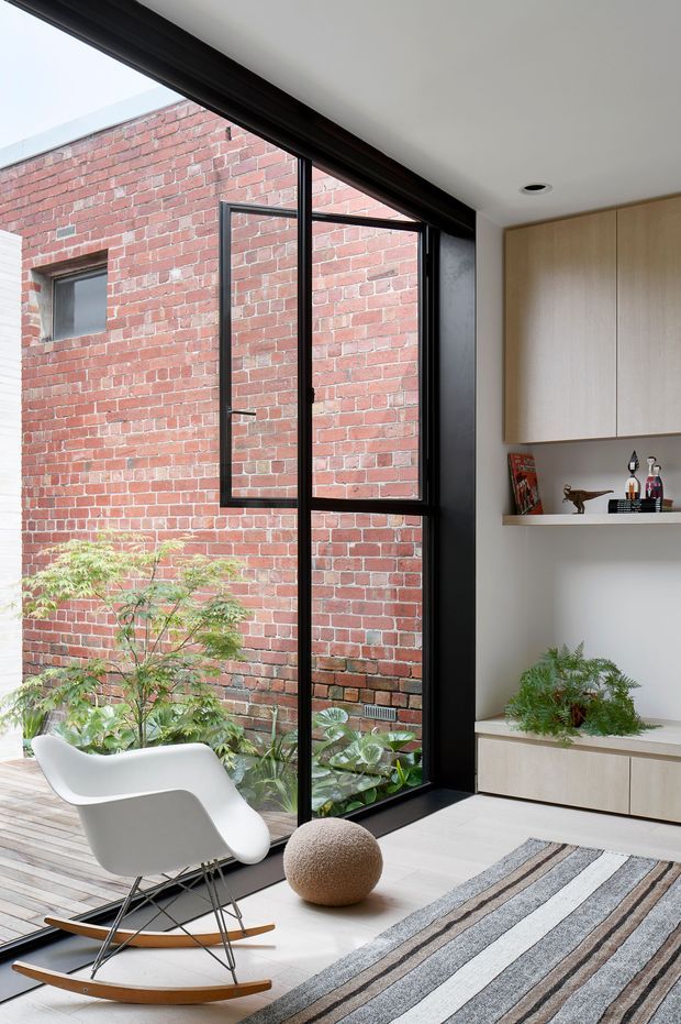 A bright, contemporary interior corner with full-height black-framed glazing overlooking a planted brick courtyard. The room includes a white sculptural rocking lounge chair, a textured round ottoman, a striped area rug, pale flooring, and built-in light-wood cabinetry with open display shelving and greenery. The exposed red brick courtyard wall is a strong visual feature.