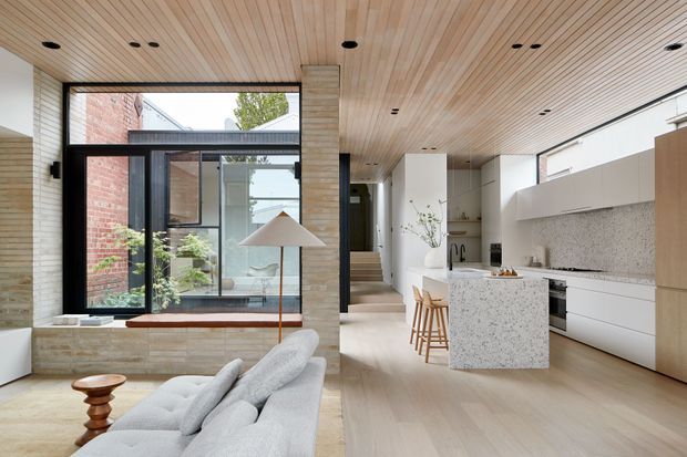 A bright, open-plan contemporary interior combining a living area and kitchen. The room features a pale timber slatted ceiling, light timber flooring, a white kitchen with a terrazzo-look island and matching splashback, pale upholstered seating, black-framed glazing, and exposed brick visible around an internal courtyard or glazed opening. A compact dining or bar area sits beside the kitchen, with stairs leading toward the rear of the home.
