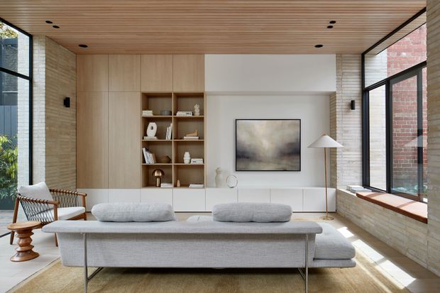 A bright, refined contemporary living room with a low-profile light grey sectional sofa facing a built-in media and display wall. The room features pale timber cabinetry and open shelving, a warm timber slatted ceiling, light textured masonry walls, black-framed floor-to-ceiling glazing, and a neutral abstract artwork. Natural light fills the space, creating an airy, calm atmosphere.