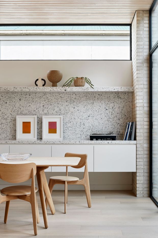 A refined dining area with a light timber dining table and chairs in front of a built-in white sideboard. The sideboard wall is finished in grey terrazzo, with open shelving displaying framed artwork, ceramics, books and a record player. A pale timber slatted ceiling, exposed light brickwork and full-height glazing create a bright, contemporary interior.
