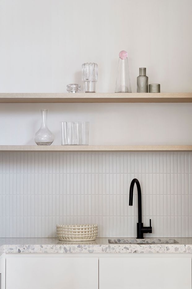 A bright, minimalist kitchen vignette featuring open pale-wood shelving displaying glassware and decorative bottles, a white vertically fluted tile backsplash, a pale terrazzo benchtop, white lower cabinetry, and a matte black gooseneck tap over a small sink. The composition is clean and softly styled with neutral tones and subtle black contrast.