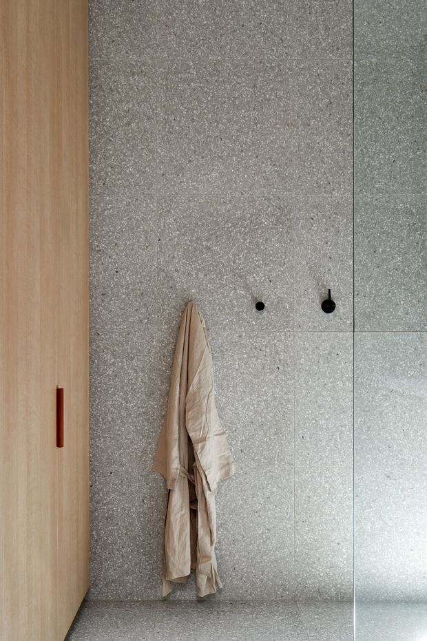 A minimalist bathroom interior featuring a full-height grey terrazzo wall and floor, a frameless glass shower screen on the right, and light timber built-in cabinetry on the left with a slim dark handle. Two black wall hooks hold a neutral robe or towel. The space has a restrained, modern material palette and clean vertical and horizontal lines.