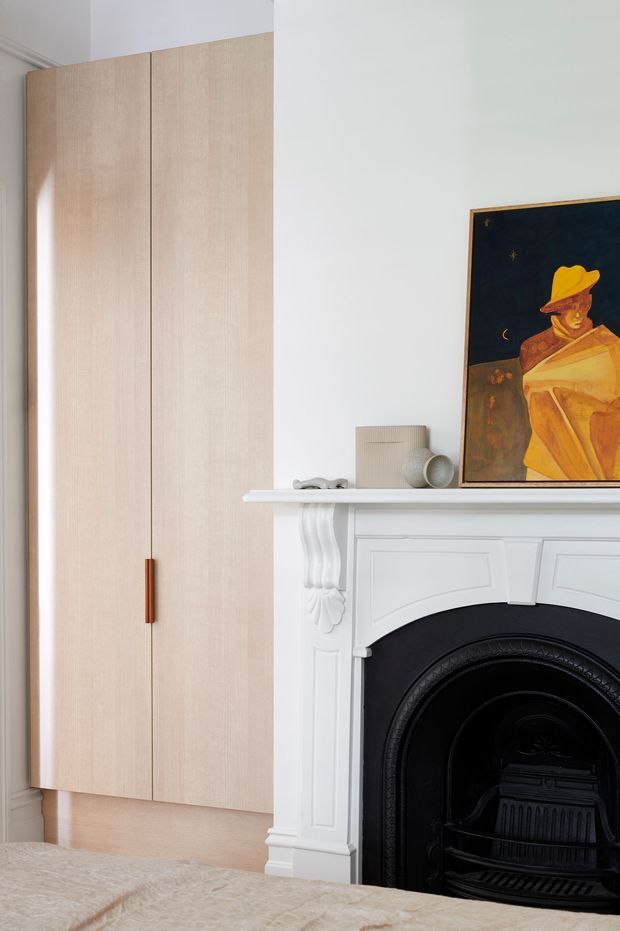 A calm, refined bedroom interior featuring a tall, pale timber built-in wardrobe with a slim copper-toned handle, beside a white ornate fireplace with black cast-iron insert. A framed artwork and small decorative objects sit on the mantel, while the edge of a bed is visible in the foreground. The room combines contemporary built-in joinery with traditional architectural detailing.