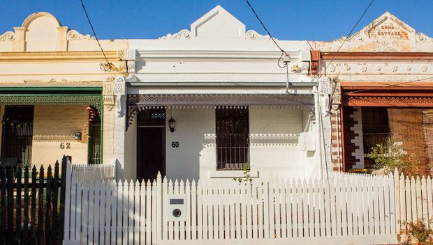 Street-facing row of narrow attached heritage terrace houses with ornate rendered façades, decorative cornices, parapets, barred windows, traditional front doors, and white picket fences. The central façade is painted white and flanked by cream and terracotta façades under a clear blue sky.