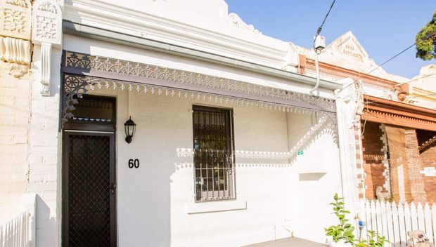 Street-facing view of a narrow heritage-style terrace or townhouse facade, painted white with ornate lacework and decorative mouldings along the parapet and awning. The facade includes a dark security door, barred window, wall-mounted lantern, house number 60, and a small white picket fence at the edge. A neighbouring exposed-brick facade is partially visible.