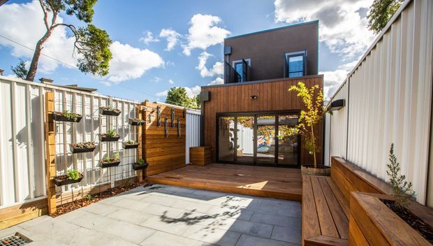 A compact contemporary courtyard or terrace enclosed by corrugated metal fencing. The space features a timber-clad two-storey structure with large sliding glass doors opening onto a timber deck, built-in timber bench seating and planter boxes, raised vertical garden planters, and large-format grey paving. Bright daylight and mature trees surround the private outdoor area.