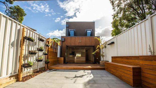 Contemporary multi-level residential exterior viewed from a paved courtyard or driveway. The compact facade combines dark rendered upper walls, vertical timber cladding, a large recessed garage or open ground-floor bay, and tall light-coloured boundary fencing with integrated timber planter boxes. The setting is sunny and landscaped with climbing plants and potted greenery.