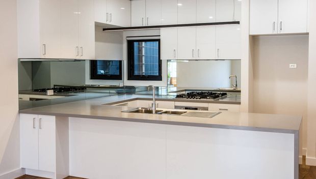 A bright, contemporary galley-style kitchen with extensive glossy white cabinetry, a large peninsula or island with an integrated double sink, stainless-steel tapware, a pale stone benchtop, mirrored splashback surfaces, and a gas cooktop beneath upper cabinets. Dark-framed windows and warm timber flooring add contrast.