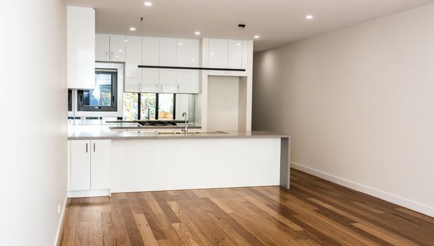 Bright contemporary open-plan kitchen with extensive glossy white cabinetry, a large white island, integrated appliances, stainless-steel sink, and warm timber flooring. The adjoining open area is unfurnished, with neutral painted walls and recessed ceiling lighting.