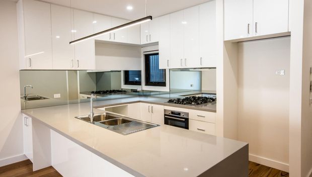 Bright contemporary white kitchen with extensive glossy flat-panel cabinetry, a large stone island with integrated double sink and drainer, mirrored splashback, gas cooktop, built-in oven, and timber flooring. A slim linear pendant provides task lighting over the island.