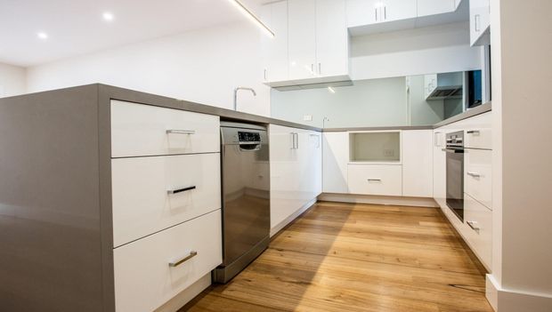 Bright contemporary kitchen with white flat-panel cabinetry, integrated appliances, a dark grey island, pale reflective splashback, and warm timber-look flooring. Recessed ceiling lighting and clean-lined joinery create a simple, modern fit-out.
