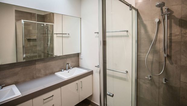 A contemporary bathroom with a white vanity featuring integrated basins, dark brown benchtop, mirrored wall cabinets, and a glass-enclosed shower. The shower walls and vanity backsplash are finished in large-format taupe-brown tiles, with chrome fixtures and fittings throughout.