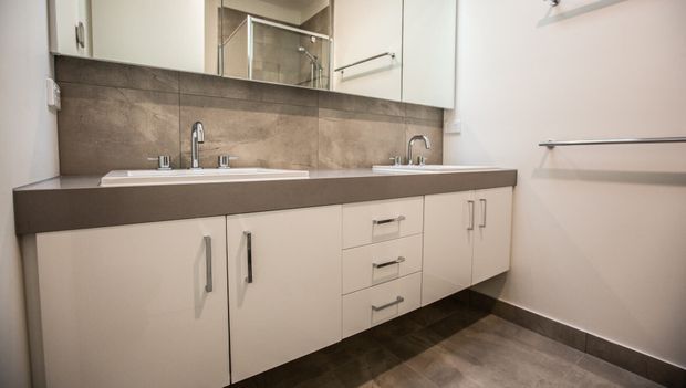 Contemporary bathroom featuring a long double vanity with white cabinetry, a dark engineered-looking benchtop, twin basins and chrome tapware. Large-format taupe-grey wall tiles form the splashback, with mirrored cabinets above and dark floor tiles below. A glass shower enclosure is visible in the reflection, and chrome towel rails are mounted on the walls.