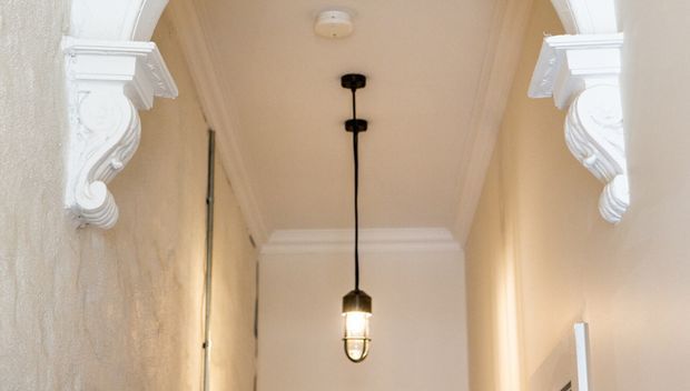 A narrow, softly lit hallway or passage with warm cream plaster walls, ornate white corbels and moulded architraves, a decorative cornice, and a hanging lantern pendant. The detailing gives the space a traditional, refined character.