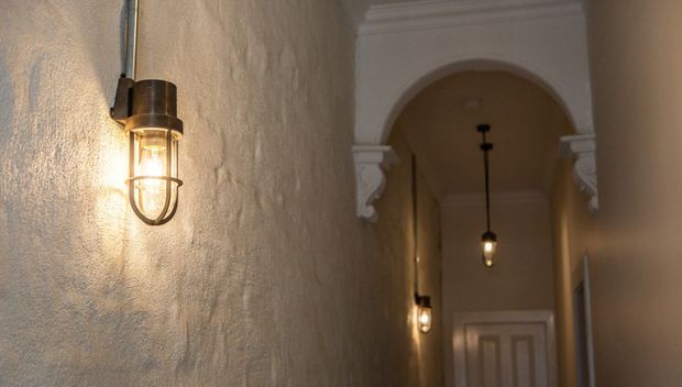 A dim, warmly lit interior hallway with textured plaster walls, a high ceiling, and a decorative arched opening framed by classical white mouldings. Industrial-style wall sconces and hanging pendant lights illuminate the corridor, with a paneled door visible at the far end.