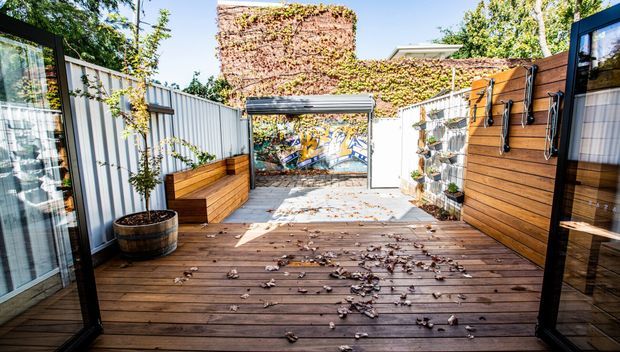 A compact urban outdoor courtyard or rooftop terrace with a dark timber deck, built-in timber bench seating, corrugated metal privacy fencing, timber-clad walls, climbing greenery, potted plants, and a small mural visible at the far end. The space is bright and enclosed, with contemporary outdoor detailing and glass doors opening onto the deck.