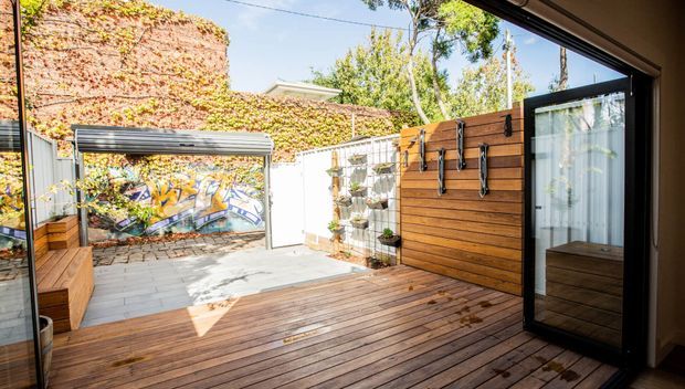 A sunlit enclosed courtyard viewed from an interior opening, featuring a raised timber deck, horizontal timber privacy wall with outdoor hanging hooks, a black-framed glazed door, rendered and ivy-covered brick walls, planted wall shelving, and a colourful mural at the far end.
