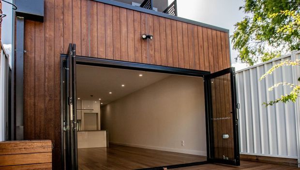 Exterior view of a compact contemporary timber-clad building or extension with large black-framed bi-fold doors opened to reveal a polished interior space with recessed ceiling lights, white walls, pale cabinetry or a counter, and dark timber flooring. A corrugated metal boundary fence and surrounding trees are visible outside.