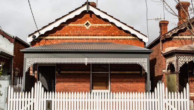 Front exterior view of a traditional single-storey red-brick house with an ornate gabled facade, decorative timber veranda fretwork, corrugated metal roof, brick chimneys, and a white picket fence.