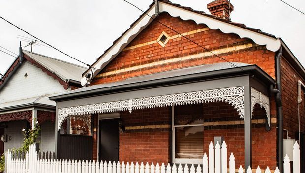 Street-facing view of a traditional single-storey red-brick house with ornate decorative gable trim, a corrugated metal roof, lacework veranda enclosure, timber picket fence, and a prominent brick chimney. A neighbouring weatherboard house is partially visible at left.
