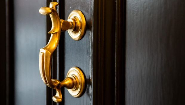 Close-up of an ornate polished brass or gold-toned pull handle mounted on a dark painted or stained wooden door, with decorative circular escutcheons and strong contrast between the warm metal and near-black door surface.