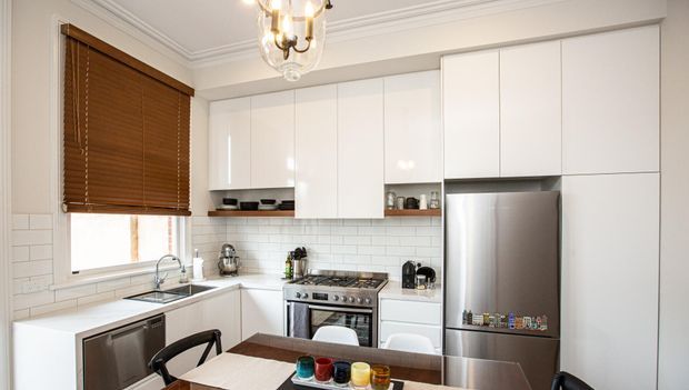 Bright, compact contemporary kitchen with white full-height cabinetry, white subway-tile splashback, stainless-steel appliances, a gas range, farmhouse-style sink, timber venetian blind, and a dining table in the foreground. Decorative ceiling mouldings and a traditional chandelier add a classic touch.
