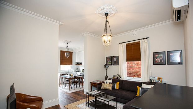 Bright traditional-style open-plan living room with a dark sofa, armchair, coffee tables, framed wall art, pendant lighting, decorative ceiling rose and cornices. A dining area and kitchen are visible through the opening, while a window with white curtains and timber blind reveals a brick wall outside.