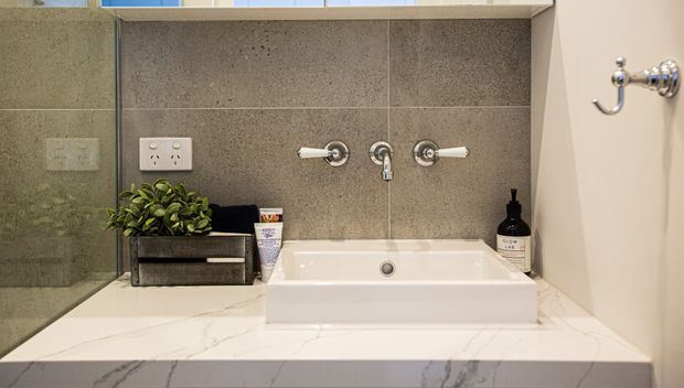 Contemporary bathroom vanity featuring a white rectangular vessel basin set on a white marble-look benchtop, with grey stone-look wall tiles, polished chrome wall-mounted tapware, a mirrored side panel, and a small built-in planter and toiletries.
