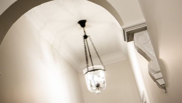 Close-up interior architectural detail of a softly lit hallway or entry passage, featuring a rounded plaster archway, ornate white decorative wall moulding or corbel, and a hanging glass pendant light. The image has a refined, traditional character with a warm neutral palette and shallow depth of field.