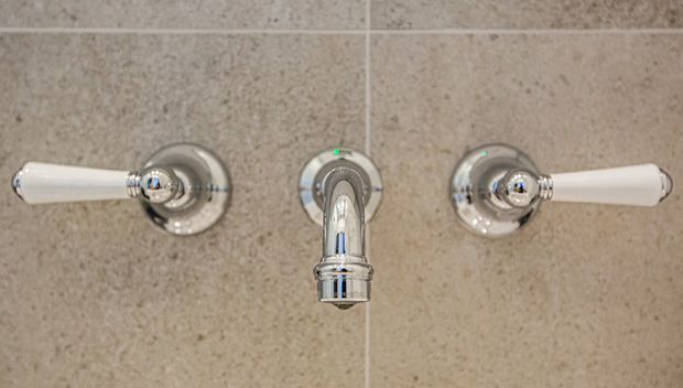 Close-up of a traditional-style wall-mounted bathroom basin faucet set, with polished chrome cross handles and central spout against beige stone-look tiled wall.