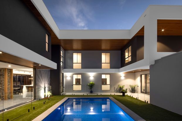 Contemporary multi-storey courtyard residence viewed from the pool, with crisp white rendered volumes, dark charcoal walls, warm timber soffits, extensive glazing, and a rectangular blue swimming pool surrounded by narrow lawns and paving. The building forms a private U-shaped courtyard with balconies and recessed outdoor areas.