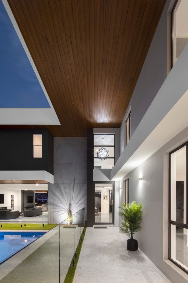 A contemporary multi-level interior courtyard or atrium with a dramatic double-height void. The space features a warm timber-lined ceiling, exposed concrete feature wall, dark and light painted walls, extensive black-framed glazing, and a pale stone or concrete floor. A glass balustrade overlooks a narrow swimming pool and a lower living area, with indoor planting adding a natural element. Recessed and wall-mounted lighting creates a refined evening atmosphere.