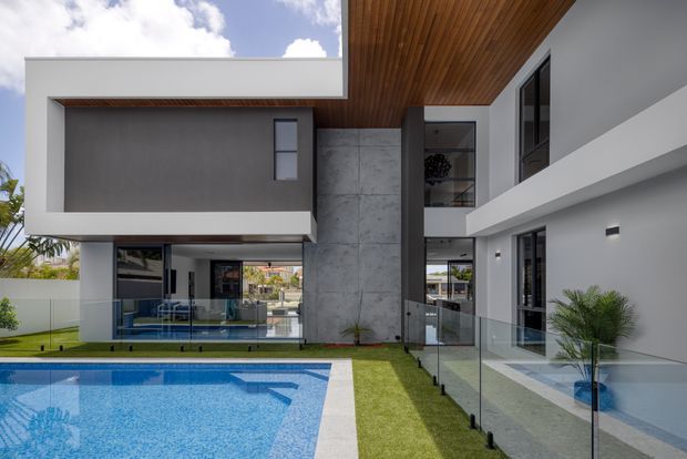 Modern multi-level residential home viewed from the pool courtyard, featuring strong geometric cantilevered volumes, a tall central concrete feature wall, extensive dark-framed glazing, glass balustrades, and a warm timber-lined soffit. A blue swimming pool and landscaped lawn occupy the foreground, with interior spaces visible through the glazing.