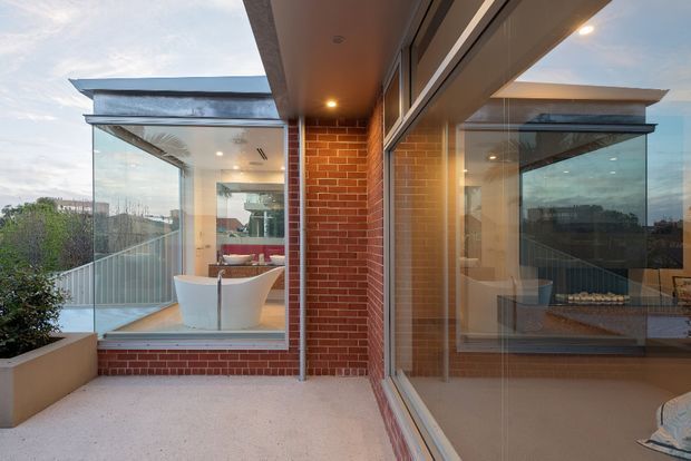 A contemporary rooftop terrace or balcony enclosed by extensive floor-to-ceiling glazing. Red brick walls and warm-toned soffits frame views into a bright bathroom with a freestanding white bathtub, while glass balustrades and neighboring urban rooftops are visible beyond. The space has a refined indoor-outdoor connection and strong linear architectural detailing.