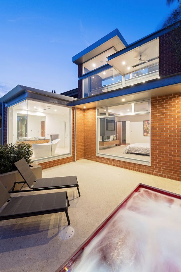 Evening exterior view of a contemporary multi-level brick residence arranged around a private courtyard. The courtyard has pale hard paving, dark outdoor loungers, extensive glazing, and a partially visible illuminated spa or pool in the foreground. Floor-to-ceiling windows reveal bright interior spaces, including a bedroom and bathroom, while an upper-level balcony with glass balustrades and a ceiling fan overlooks the courtyard.